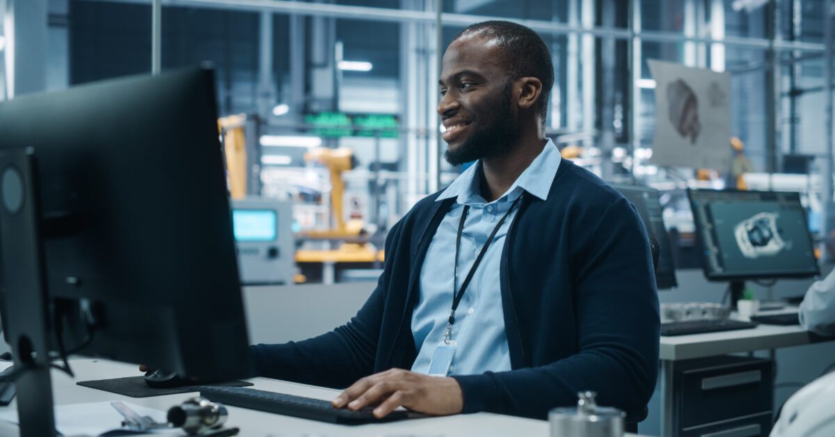 Car Factory Office: Portrait of Confident Black Male Chief Engineer Working on Desktop Computer. Professional Technician in Automated Robot Arm Assembly Line Manufacturing High-Tech Electric Vehicles